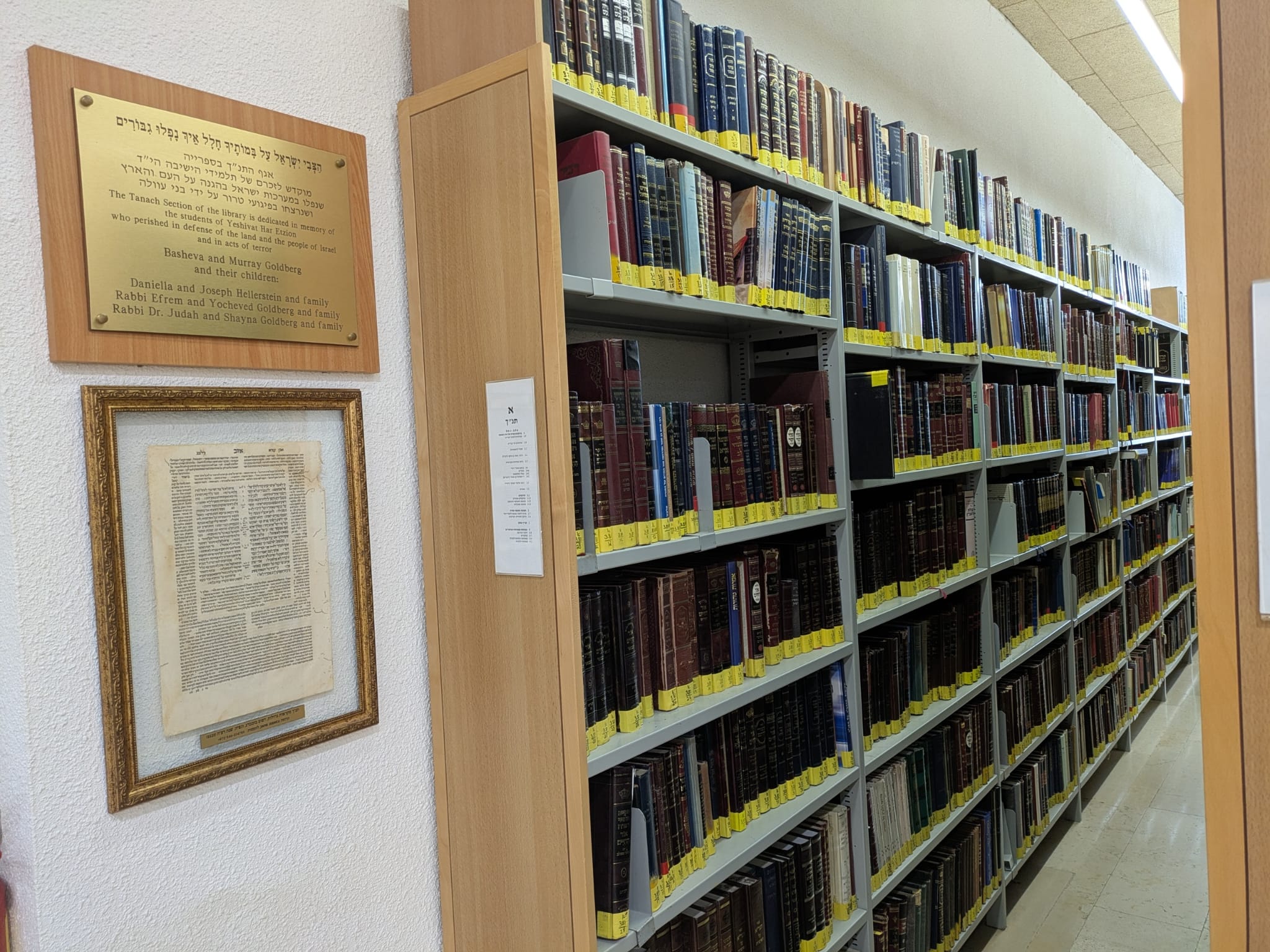 דף בומברג ממוסגר תלוי על קיר בספריית מכון הר עציון, לצד שלט הקדשה, ולידו ספריית תנ"ך